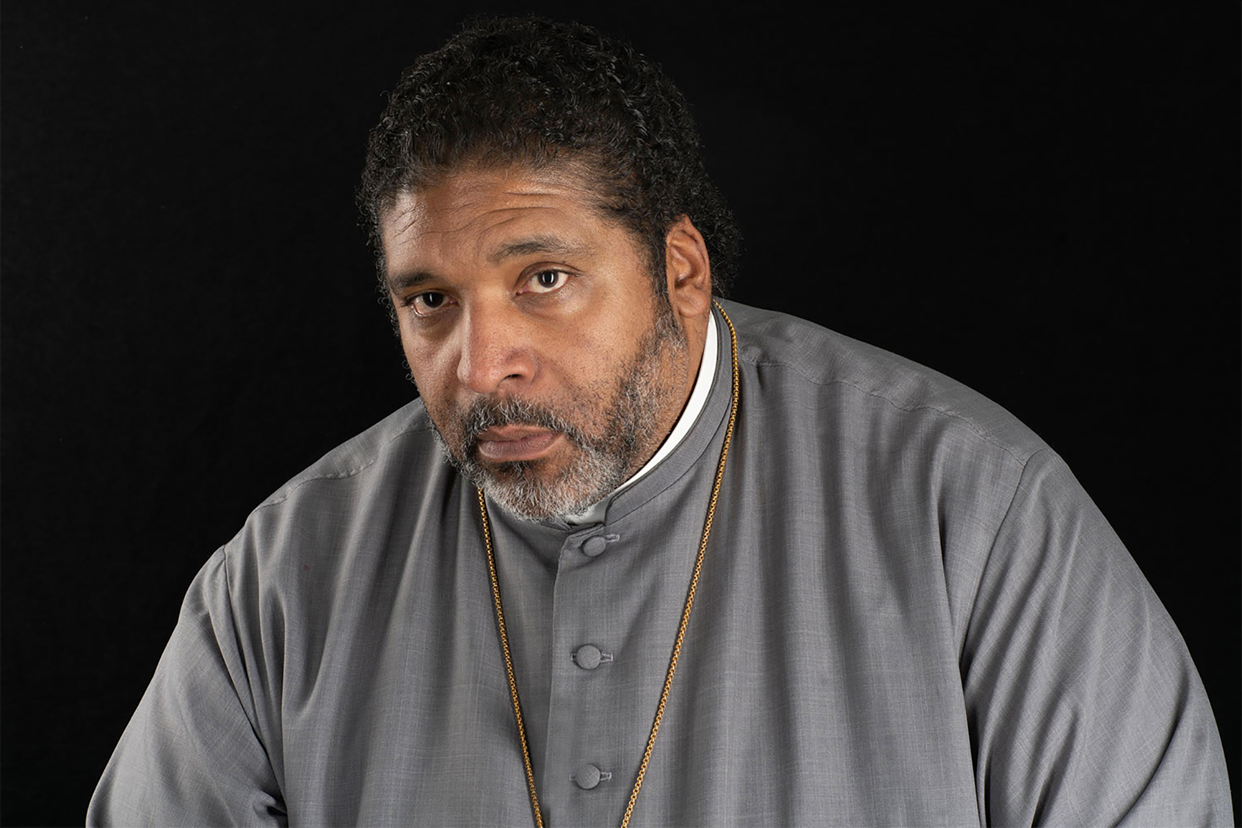 Rev Barber is staring into the camera. Wearing a gray shirt, priest collar, and a gold necklace